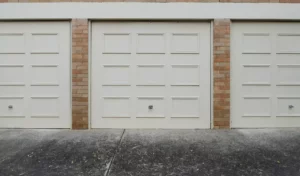 A picture of 3 cream garage doors with wood panels on blonde brick with one of the most most common garage door problems, being stuck closed or very slow.
