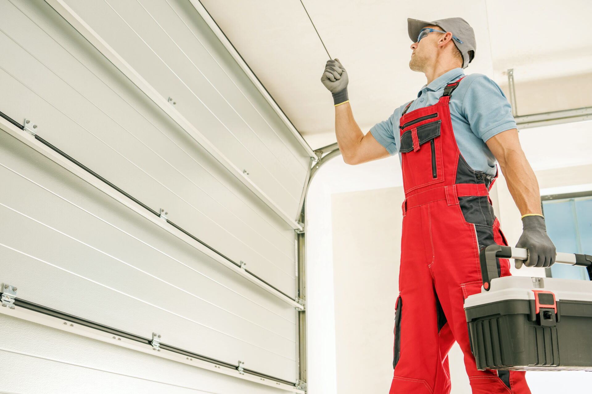 A man in orange worker's overalls stands inside an empty garage. One hand holds a tool box, and the other pulls the garage door's emergency pull string.